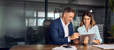 Team of diverse partners sitting at table mature Latin business man and European business woman discussing project on tablet in office. Two colleagues of professional business people working together.